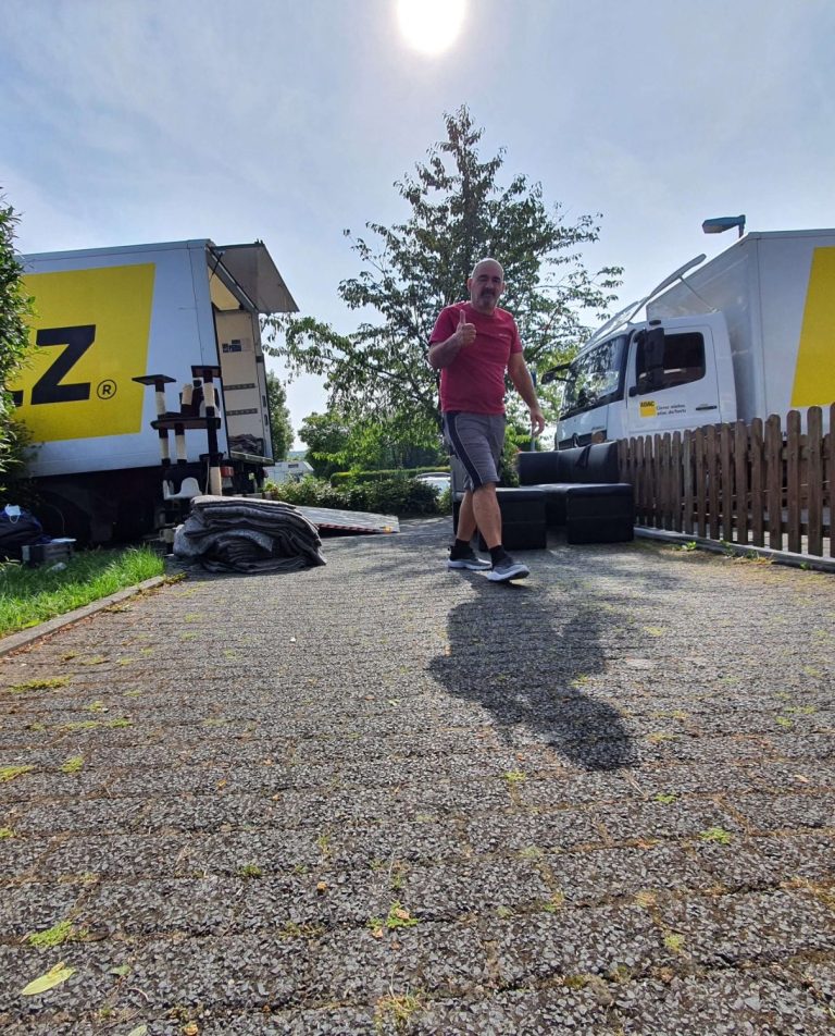 Person in rotem Shirt steht auf einer Auffahrt zwischen zwei Lieferwagen und zeigt Daumen nach oben